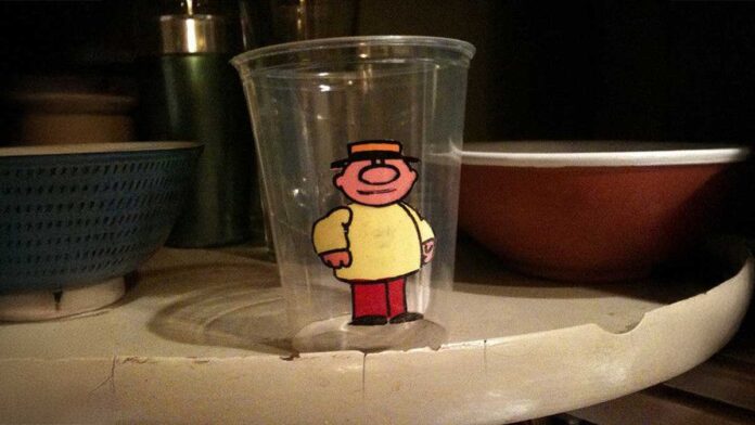 A clear plastic cup features a cartoon character with a hat and round nose, wearing a yellow shirt and red pants. The cup is placed on a slightly worn, curved shelf, with a blue bowl and other items in the background.