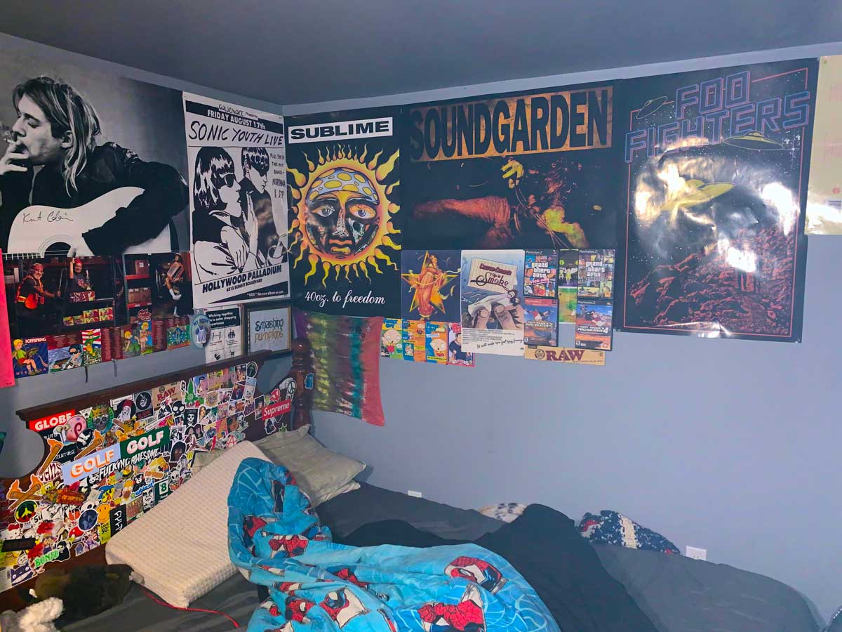 A bedroom with band posters for Nirvana, Sonic Youth, Sublime, Soundgarden, and Foo Fighters on the wall above a bed. The bed has a colorful comforter and pillows, with stickers and photos decorating the room.