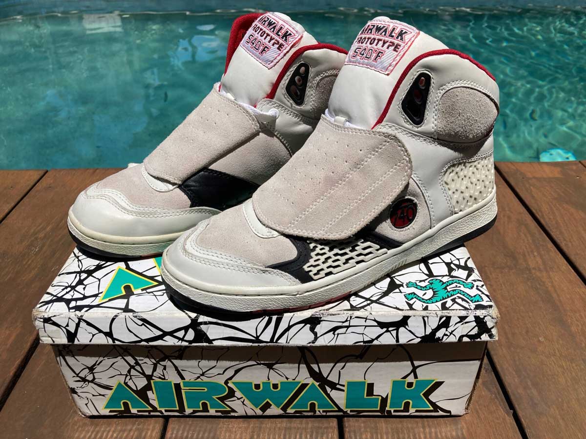 A pair of vintage white and gray Airwalk basketball sneakers with red accents displayed on top of a colorful Airwalk-branded shoe box, set on a wooden surface by a swimming pool.