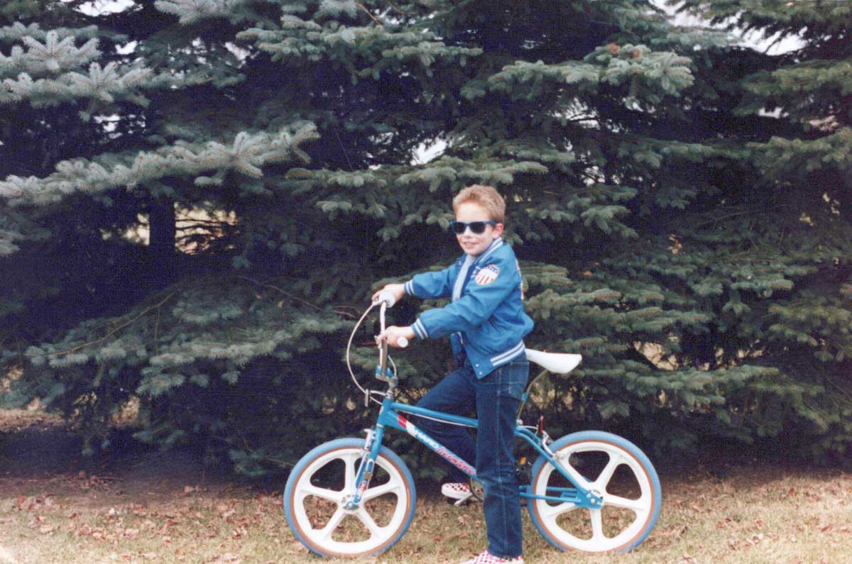 A young boy in a blue jacket and sunglasses poses on a BMX bike with white wheels in front of large evergreen trees on a grassy area.