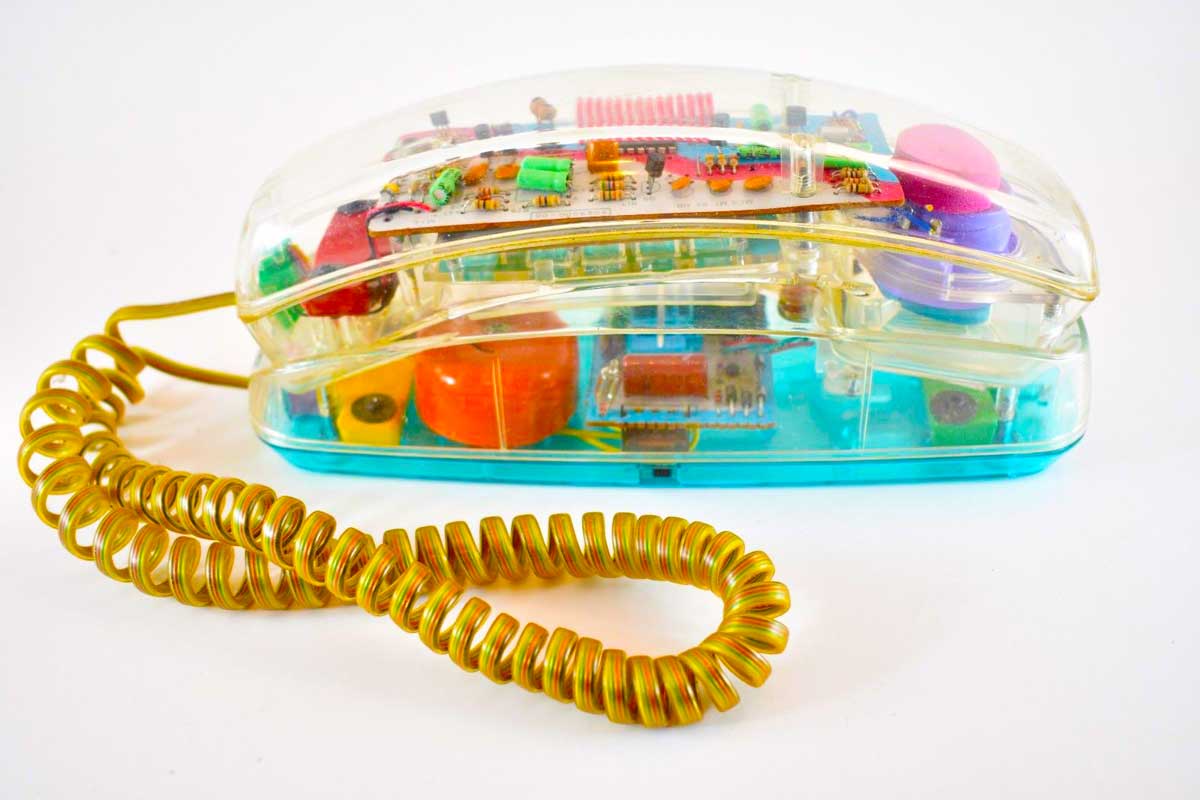 A vintage transparent landline telephone with visible internal electronic components and a coiled yellow cord, set against a plain white background.