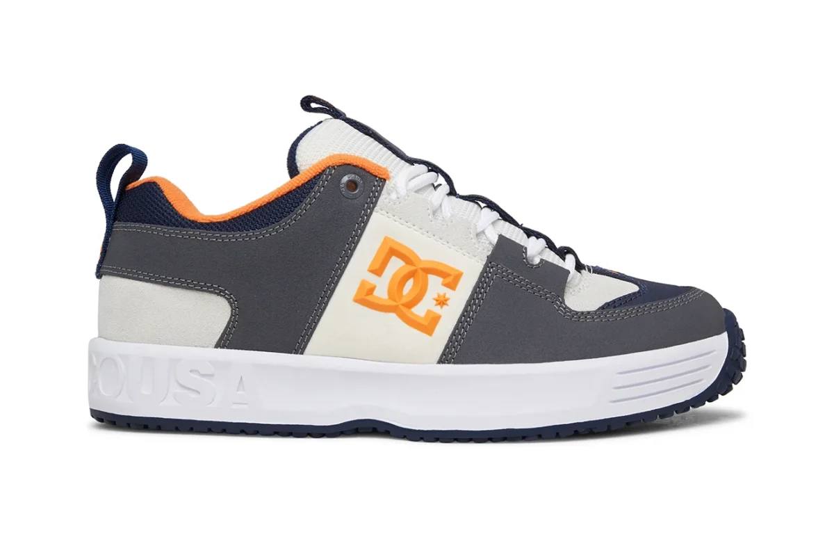A grey, white, and navy skate shoe with orange accents and a white sole. The side features a large orange DC logo, and the heel has the embossed text “USA.” The shoe has black and white laces and an orange inner lining.