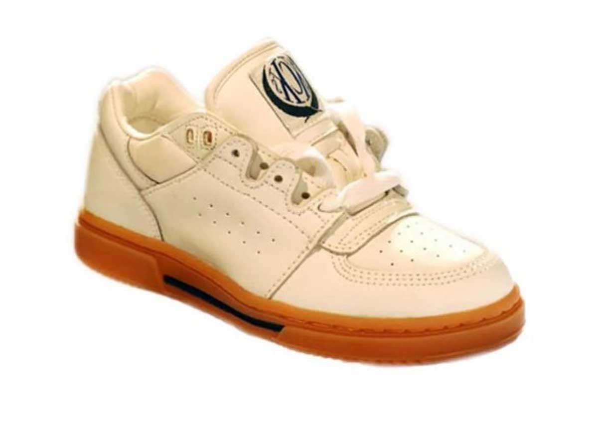 A cream-colored sneaker with a gum sole, white laces, perforated toe, and a round black-and-white logo on the tongue, displayed on a plain white background.