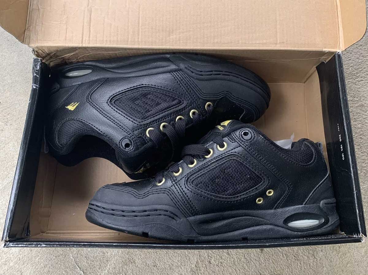 A pair of black athletic shoes with gold eyelets and accents are placed side by side inside an open cardboard shoe box. The shoes have thick soles and a mesh-and-leather design.