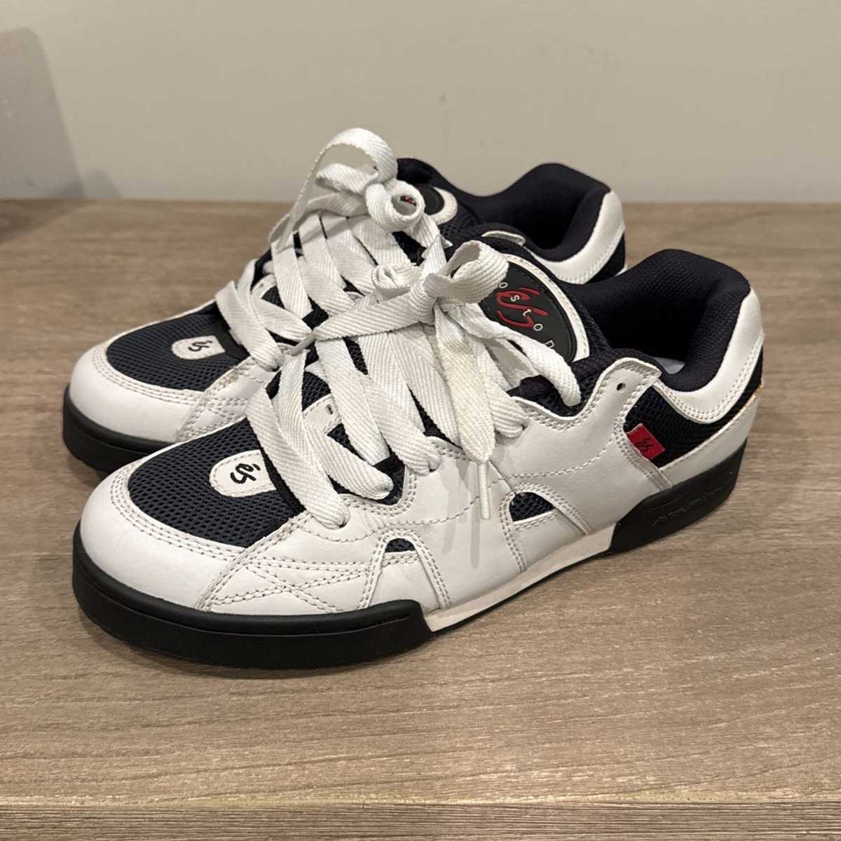 A pair of white and black skate shoes with thick white laces, black mesh sections, and red and black accents, placed on a wooden surface.