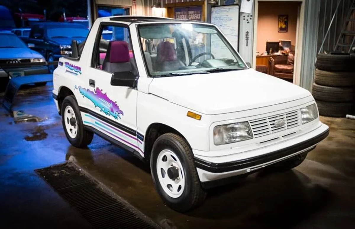 A white Suzuki Sidekick convertible with colorful retro graphics on the side, parked indoors in a garage or workshop with other vehicles and tools visible in the background.
