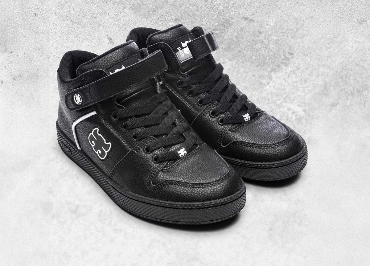 A pair of black high-top sneakers with black laces, white stitching, a Velcro strap, and white logo designs on the side and tongue, placed on a light gray textured surface.