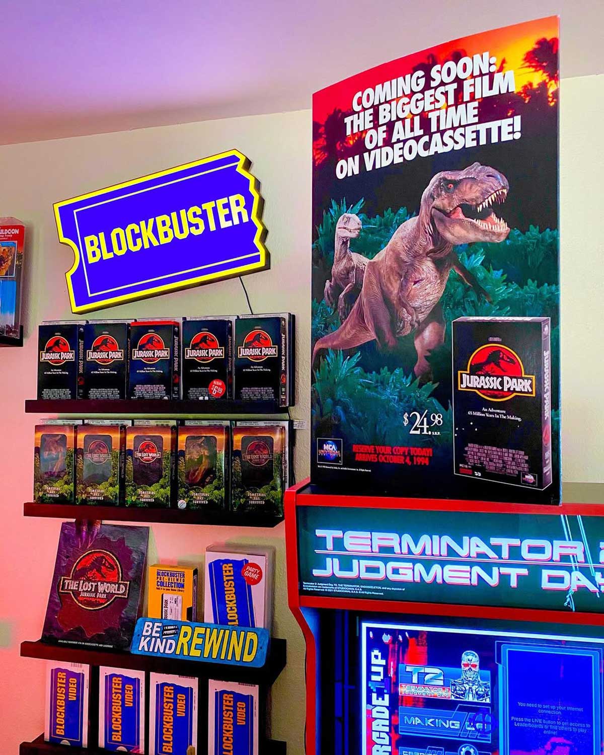 A colorful room with a Blockbuster sign, shelves of Jurassic Park VHS tapes, a large Jurassic Park poster, and a Terminator 2 arcade game, evoking 1990s video rental nostalgia.