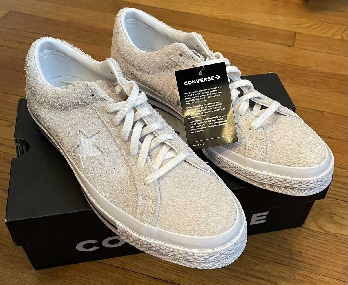 A pair of off-white Converse sneakers with white laces sits on top of a black Converse shoe box, with a black product tag attached to one shoe. The background is a wooden floor.