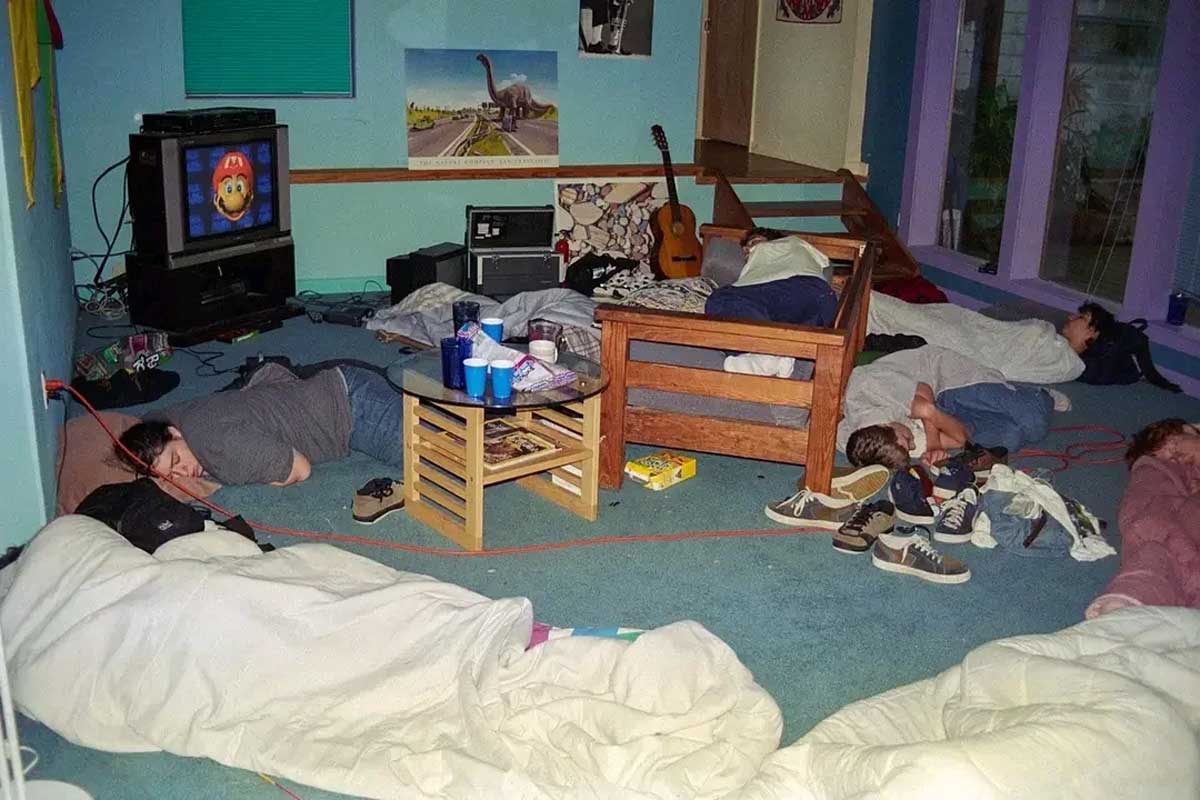 A group of people are sleeping in sleeping bags on the floor of a cluttered living room with a TV displaying a Mario game, a guitar, shoes, and cups scattered around. It looks like a sleepover.