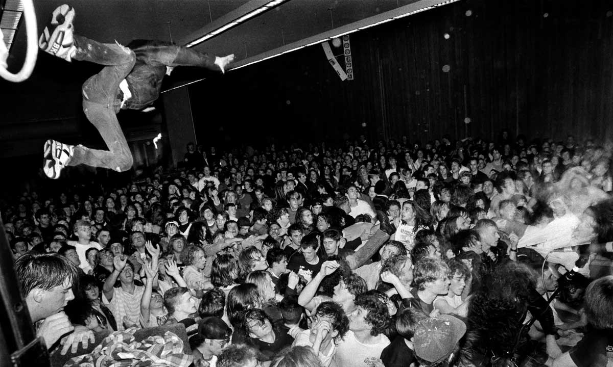 A person leaps off the stage into a packed, energetic crowd at a concert. The audience looks up, some with raised arms, capturing the chaotic and lively atmosphere of the event.
