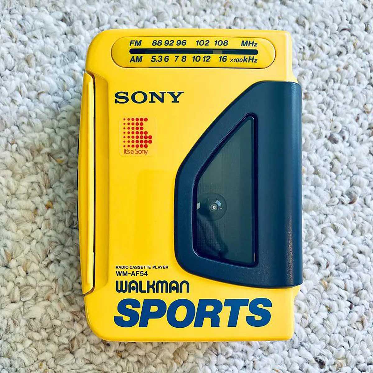 A yellow Sony Walkman Sports cassette player with blue text and a small window sits on a light-colored, textured carpet. FM and AM tuning ranges are printed on the top.