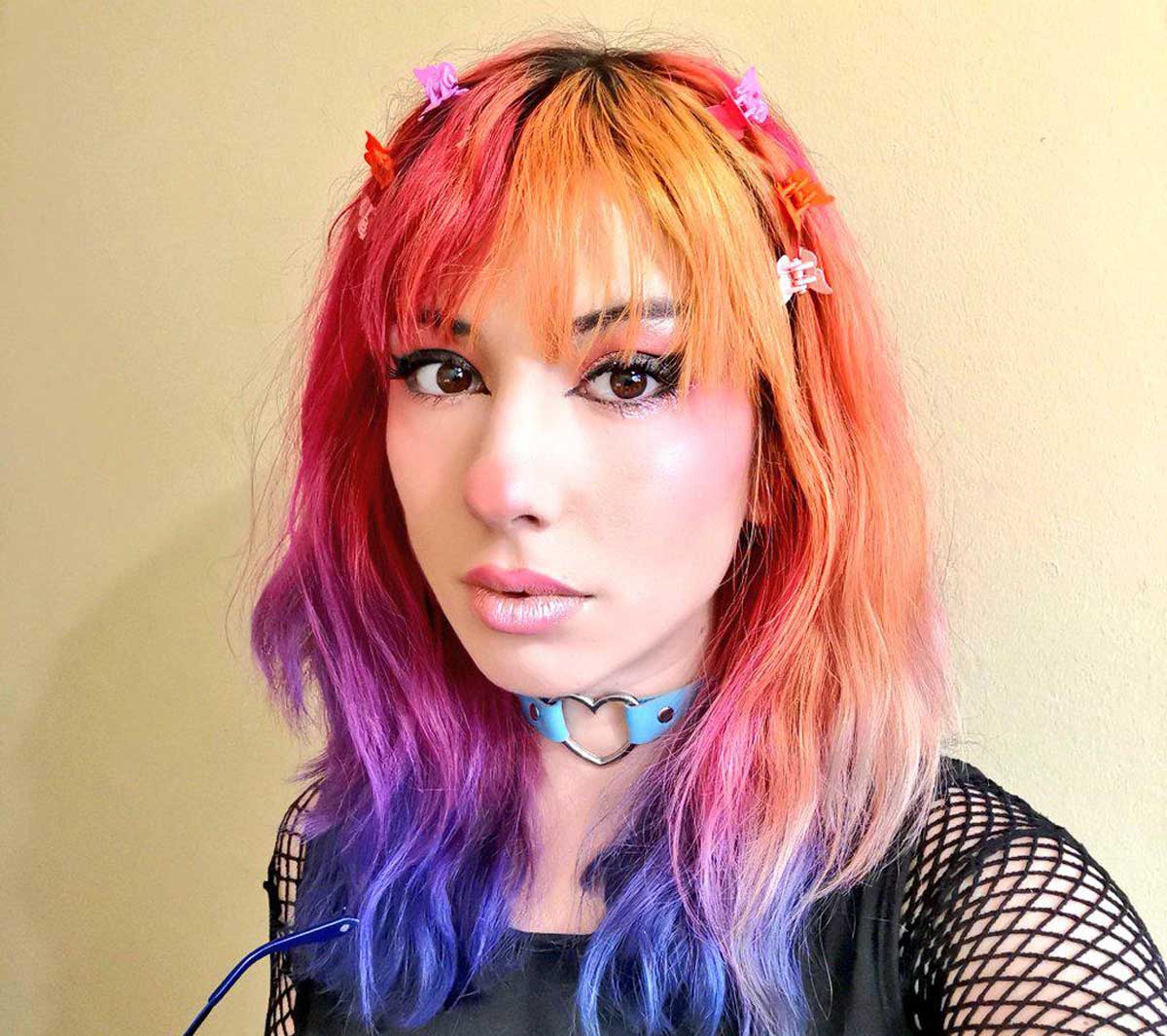 A person with orange, pink, and purple hair wears colorful clips and a light blue heart-shaped choker. They have winged eyeliner, glossy lips, and a black mesh top, standing against a plain light yellow wall.