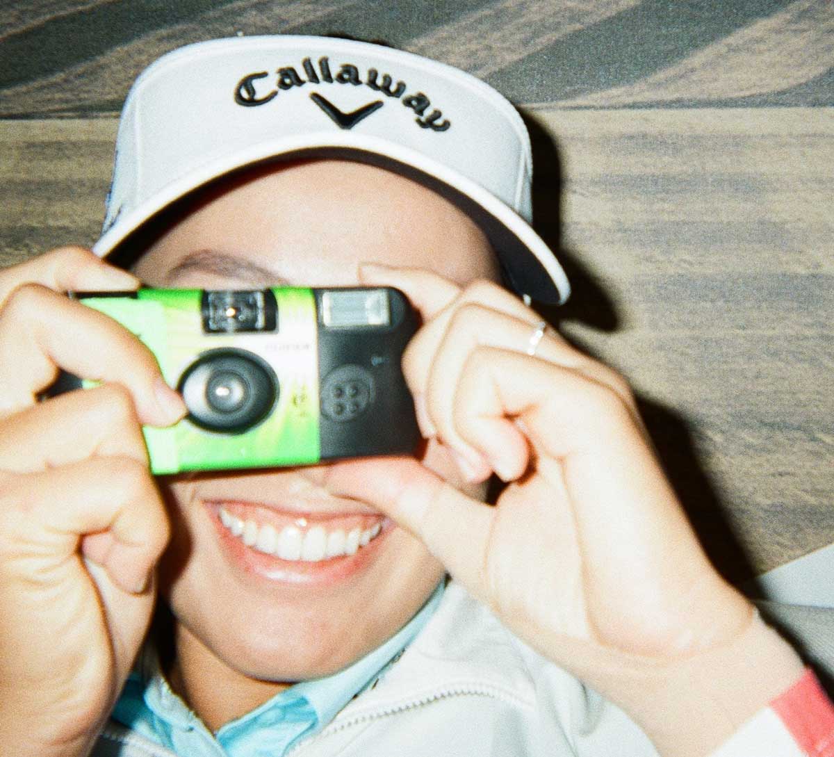 A person wearing a white Callaway cap smiles while holding a green disposable camera up to their face, as if taking a photo.
