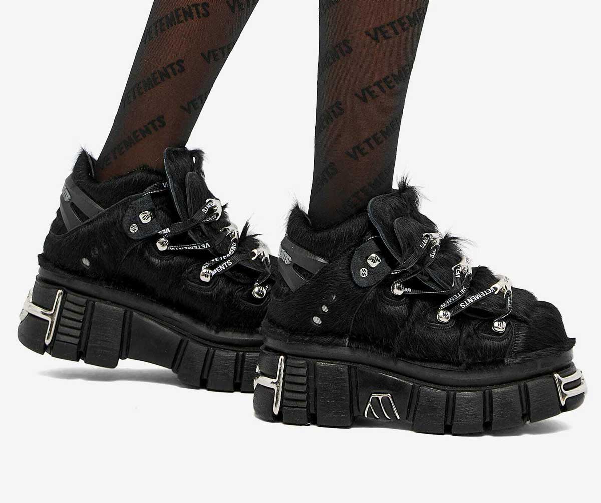 A person wears chunky black platform shoes with faux fur, metallic details, and thick lug soles. Their tights are black with the word VETEMENTS repeatedly printed in all caps.