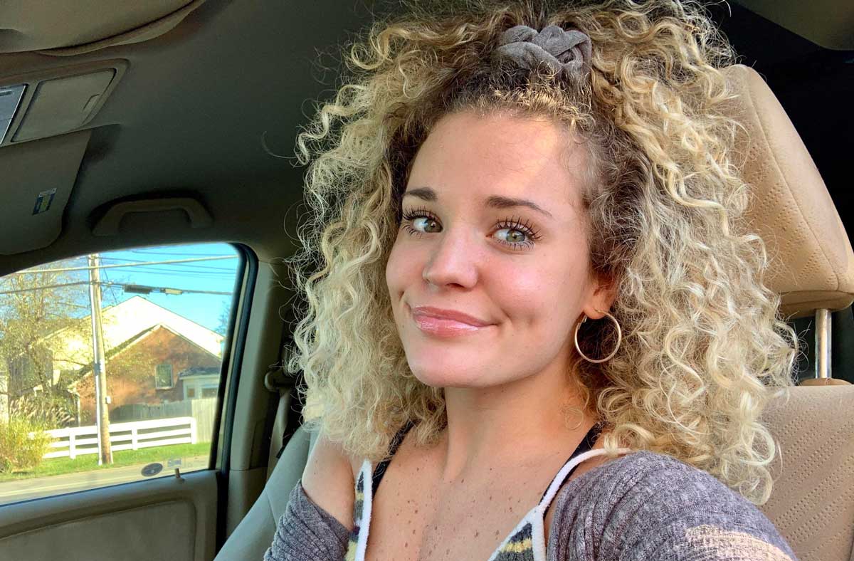 A woman with curly blonde hair and hoop earrings smiles while sitting in a car. She wears a gray top and a hair scrunchie, with a house and fence visible outside the window in the background.