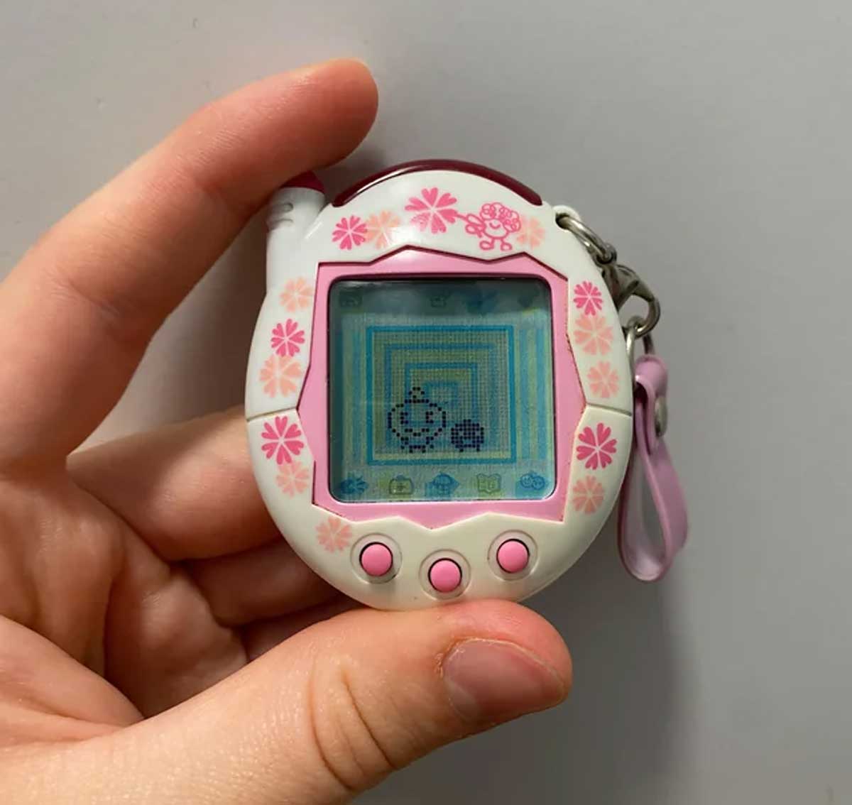 A hand holds a white and pink Tamagotchi toy with a floral design. The screen displays a pixelated virtual pet, and the device has three pink buttons and a pink strap attached.