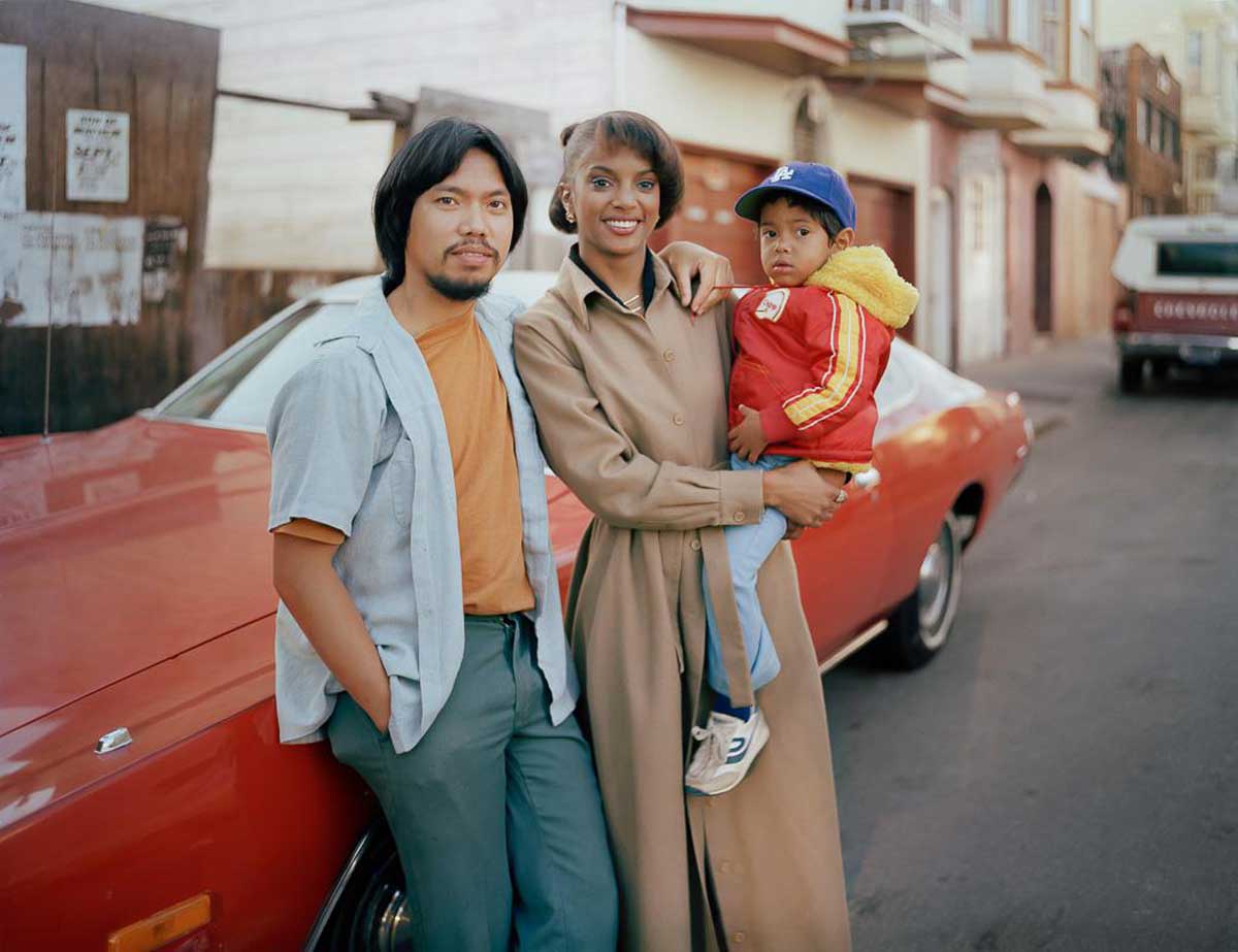 A man leans against a vintage red car while a woman in a tan coat holds a young child in a red jacket and blue cap. They are standing together on a residential street with houses visible in the background.