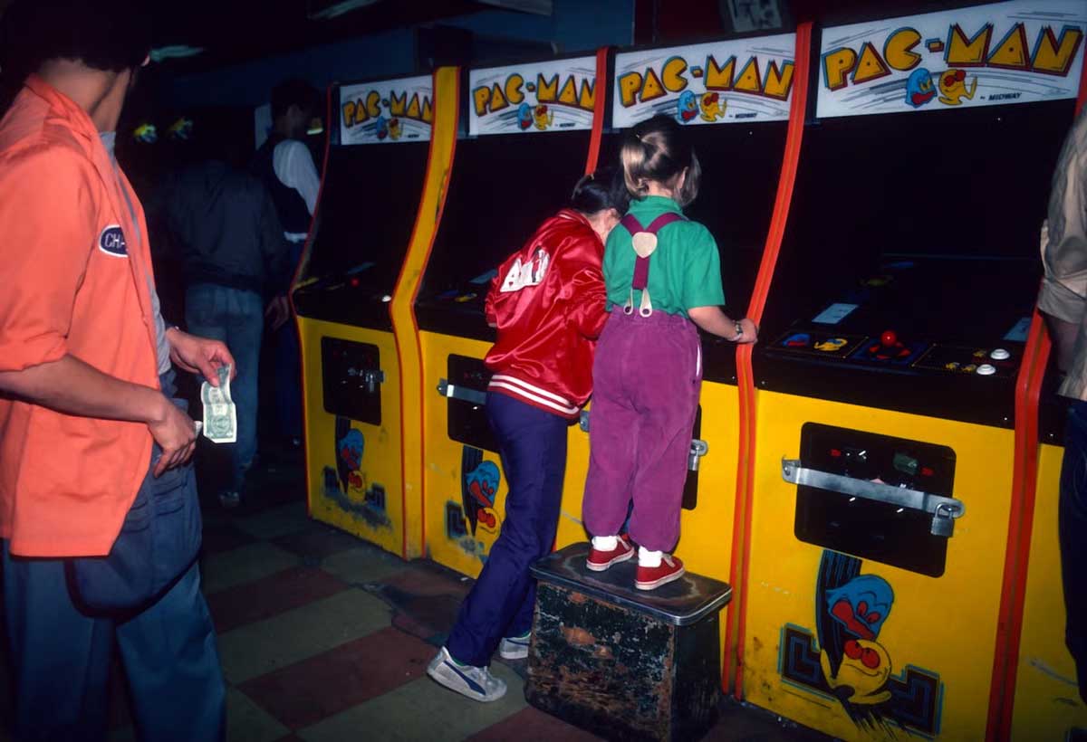 Two children play Pac-Man arcade games; one stands on a stool to reach the controls. Other people are nearby, including a person holding money. The arcade has a dimly lit, retro atmosphere.