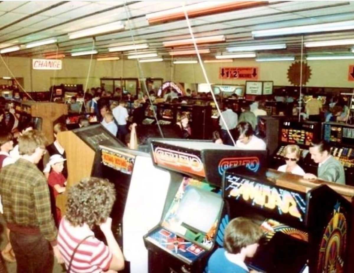 A crowded arcade packed with people playing classic arcade machines; rows of games line the room under fluorescent lights, and groups of people gather to watch or play. Signs for “Change” and “12” hang overhead.