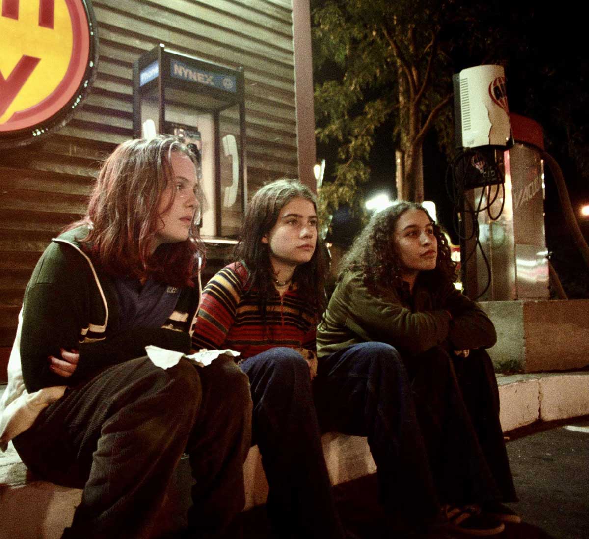 Three young people sit on a curb at night near a payphone and a gas pump, looking pensive. They are dressed casually, with streetlights illuminating the scene.