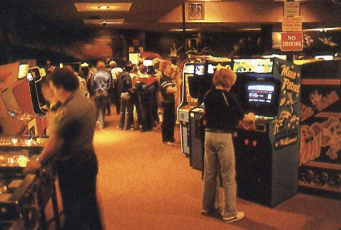 A group of people play arcade and pinball machines in a dimly-lit, crowded 1980s-style arcade. Some players face game screens, and others walk through aisles lined with colorful game cabinets.