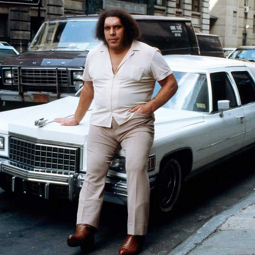 Andre the Giant with curly hair, wearing a light-colored short-sleeve shirt and pants, leans against the hood of a large white car parked on a city street.