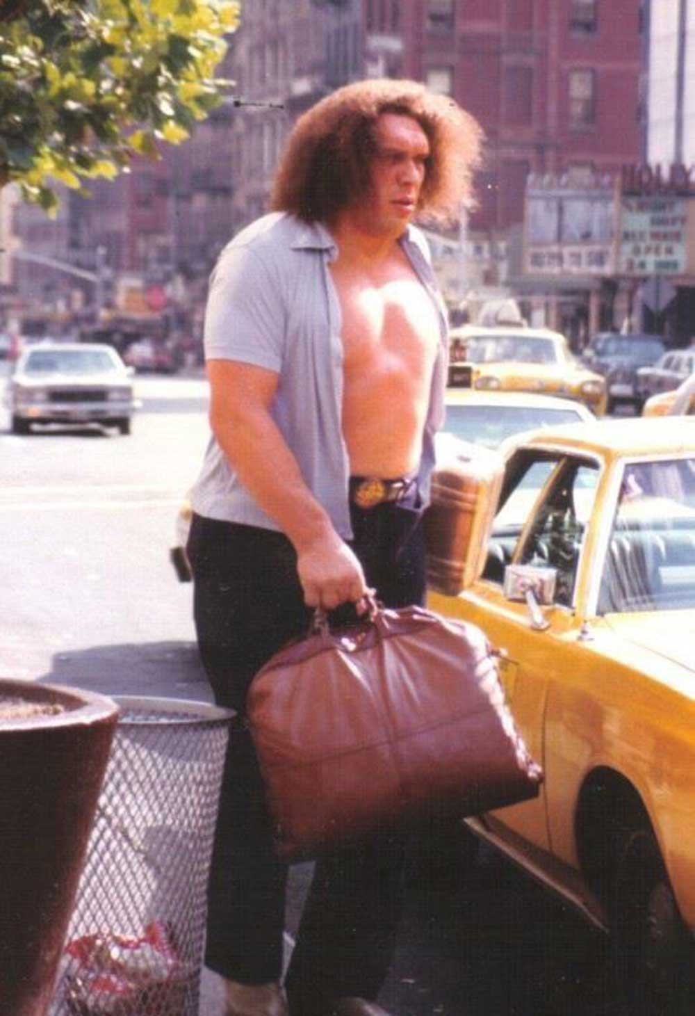 A very tall, muscular Andre the Giant with long hair, wearing an open shirt and dark pants, holds a large brown duffel bag while standing on a city sidewalk next to a yellow taxi. Urban buildings and traffic are visible in the background.