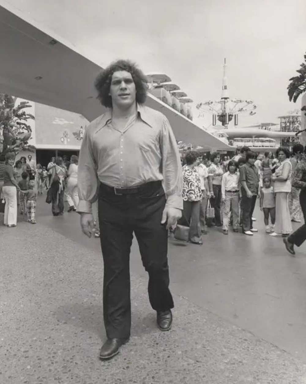 Andre the Giant with curly hair walks towards the camera in a crowded outdoor area, dressed in a button-down shirt and dark pants. People in 1970s-style clothing watch him as he passes by.