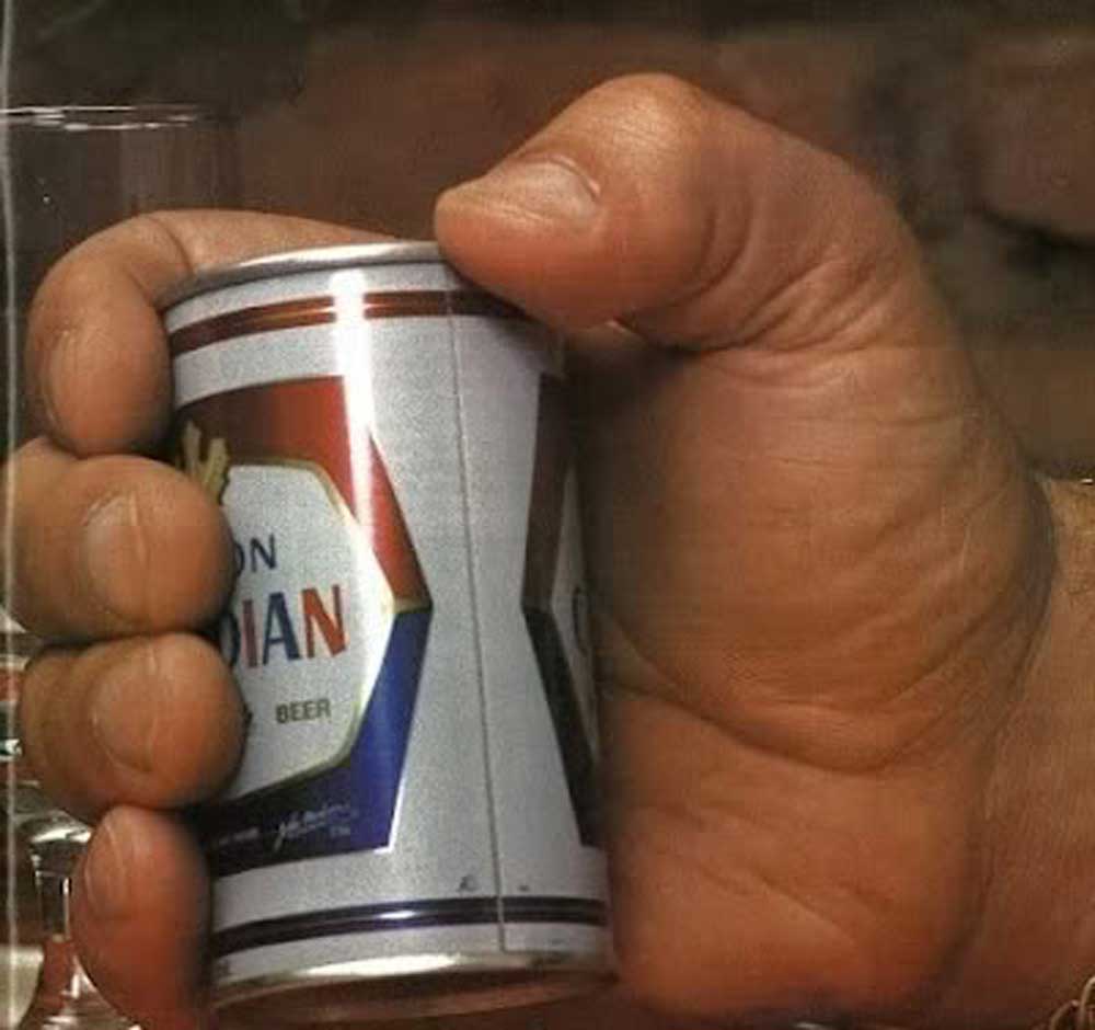 Andre the Giants very large hand holding a small can of beer, making the can appear tiny in comparison to the hand’s size. A glass is partially visible in the background.