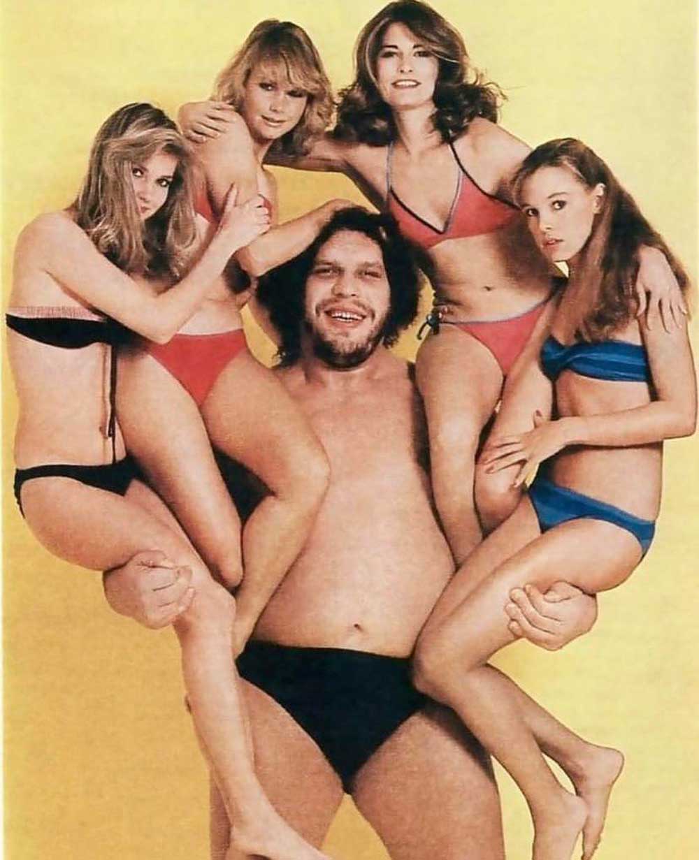 Andre the Giant in black swim trunks poses, smiling, holding four women in bikinis, with another woman standing behind him, all against a yellow background.