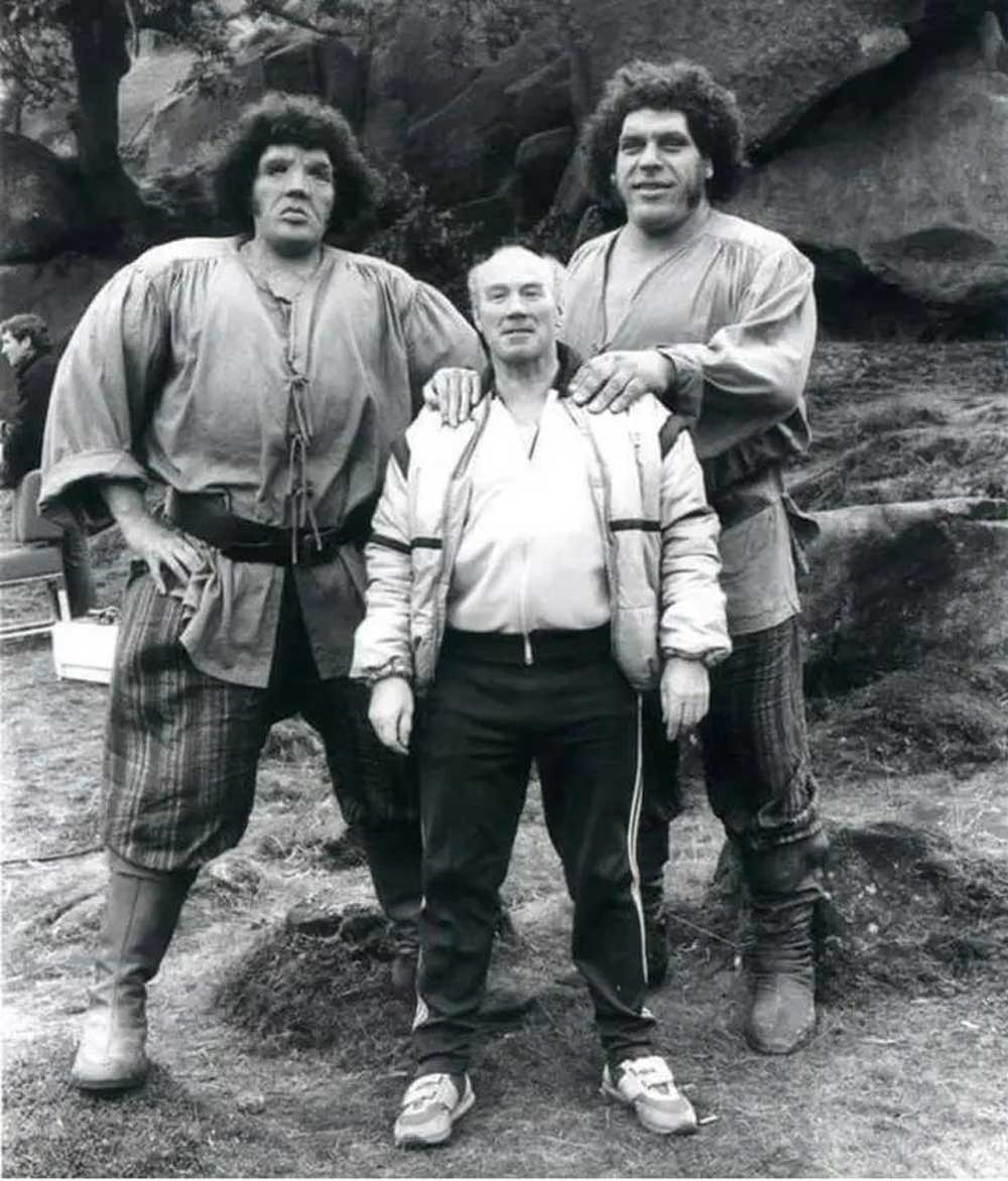 Andre the Giant and his body double in period costumes stand outdoors on either side of a much shorter man in modern clothing. Andre rests his hands on the shorter mans shoulders. All three face the camera and smile.