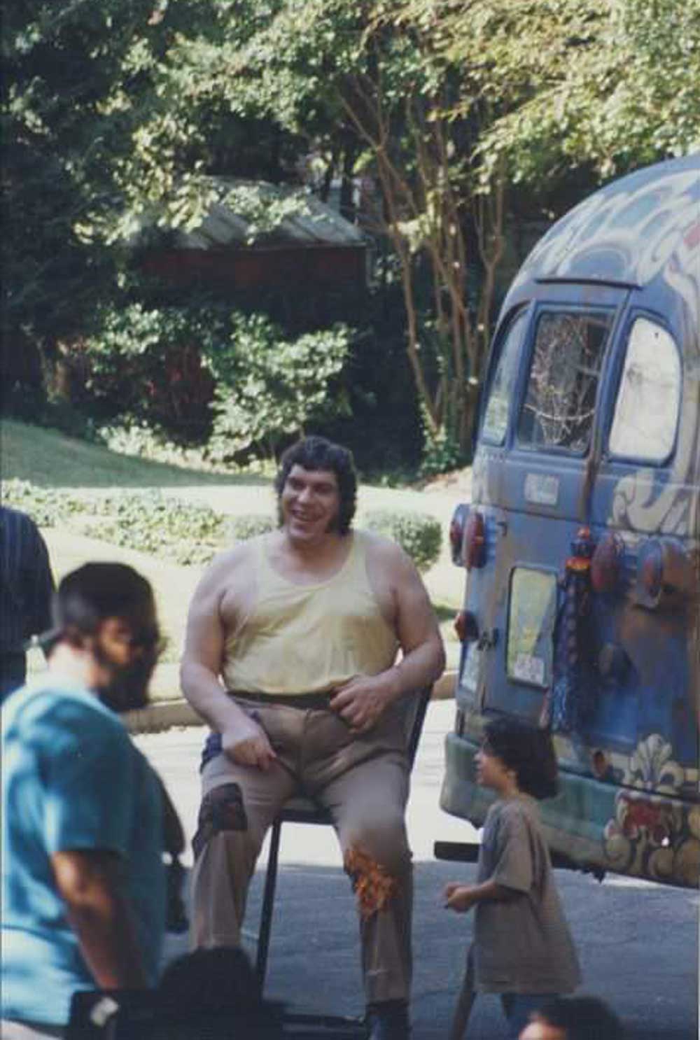A large man in a yellow tank top and brown pants sits on a stool, smiling, beside a colorful vintage van. Trees and greenery fill the background, and a child walks nearby.