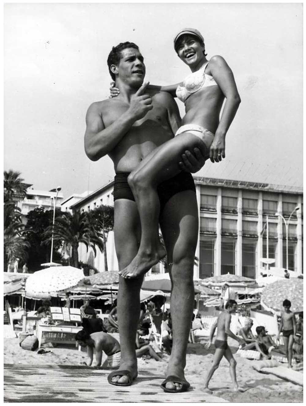A smiling Andre the Giant in swim trunks lifts a woman in a bikini at a busy beach, with sunbathers, umbrellas, palm trees, and a large building visible in the background.