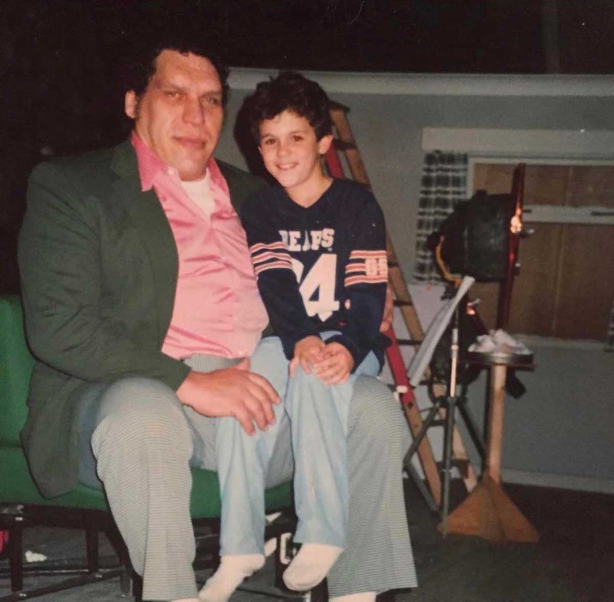 Andre the Giant in a green jacket and pink shirt sits with a young Fred Savage wearing a navy blue sports jersey on his lap. They are indoors, with a ladder and various household items visible in the background.
