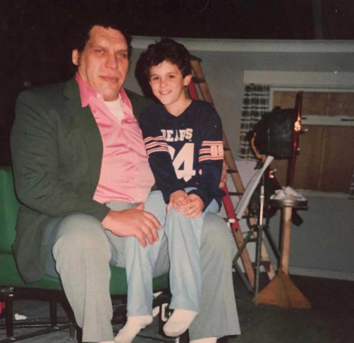 Andre the Giant in a pink shirt and green jacket sits on a bench with a smiling Fred Savage in a blue sports jersey on his lap. They are indoors, with ladders and studio equipment in the background.