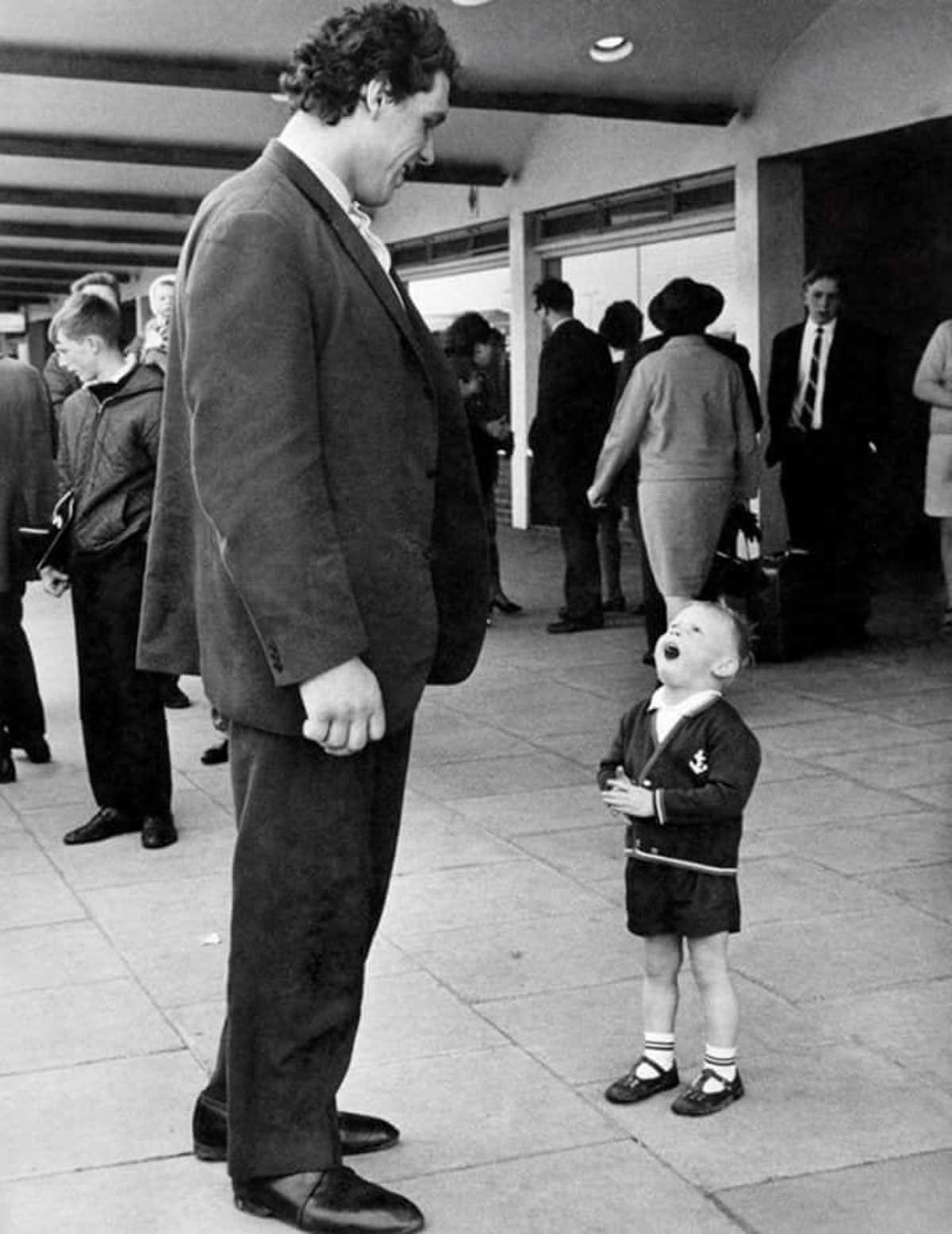 Andre the Giant smiles down at a small child looking up at him in wonder. They stand outside among a group of people, highlighting the contrast in their heights.