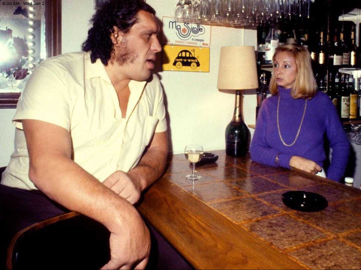 Andre the Giant in a short-sleeve shirt sits at a bar, speaking to a woman in a purple turtleneck. A drink is on the bar, and bottles line the shelves behind them. A lamp and a yellow sign are also visible.