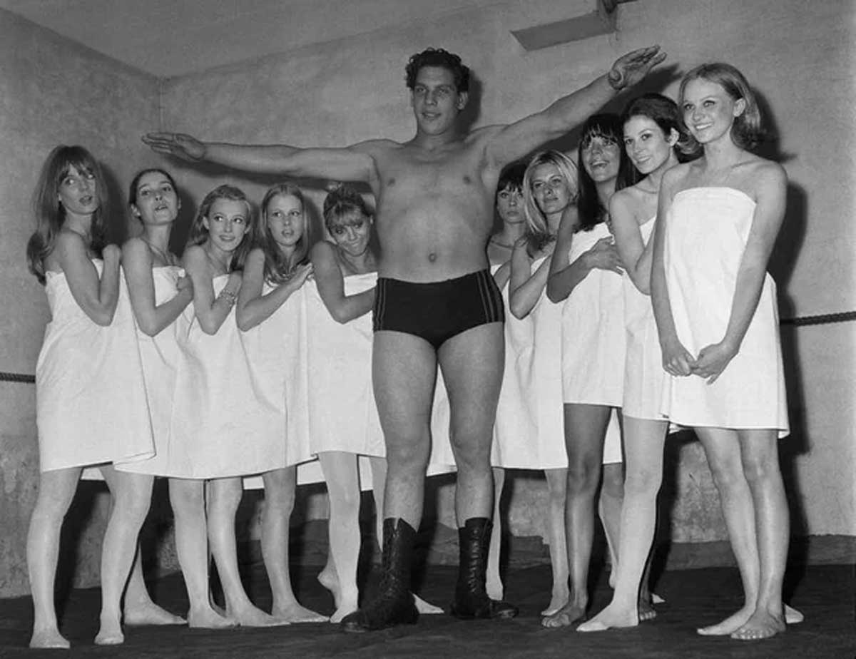 Vintage photo of Andre the Giant in black trunks and boots stands with arms outstretched, surrounded by eight women in white towels. The group poses indoors against a plain wall, with the women gathered closely around the man.