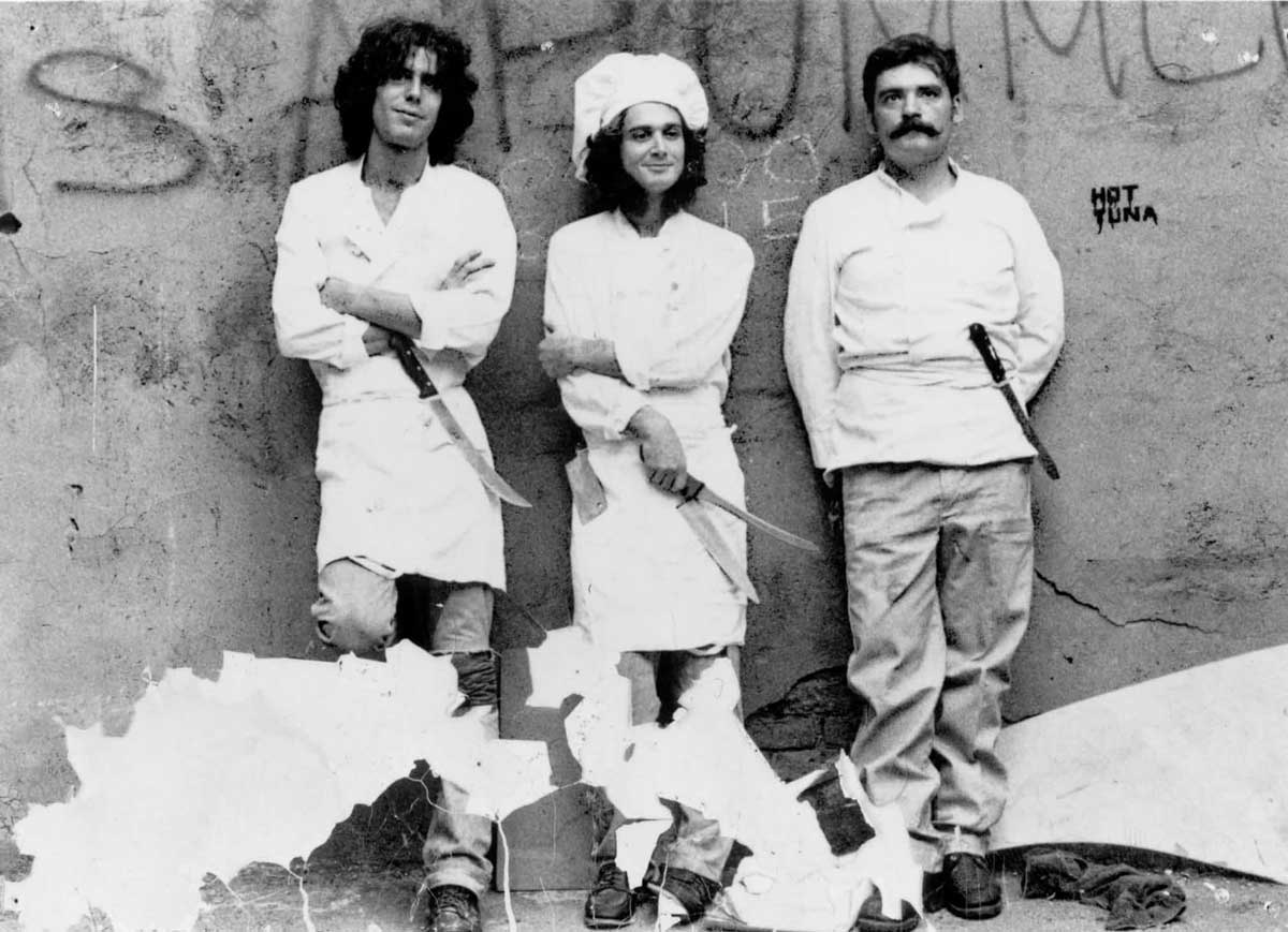 Three people wearing chef outfits stand against a worn, graffiti-marked wall. Two hold knives, one has a mustache, and all strike relaxed poses. The wall has visible peeling paint and the words HOT TUNA written on it.