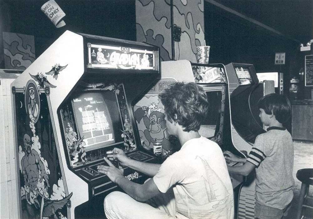 Two boys play classic arcade games at standing cabinets in a vintage arcade. The cabinets feature colorful artwork and joysticks, with soda cups placed on top. The scene has a nostalgic, 1980s atmosphere.