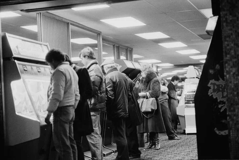 A group of people, dressed in winter clothing, stand in a row playing arcade machines in a dimly lit arcade with fluorescent ceiling lights.