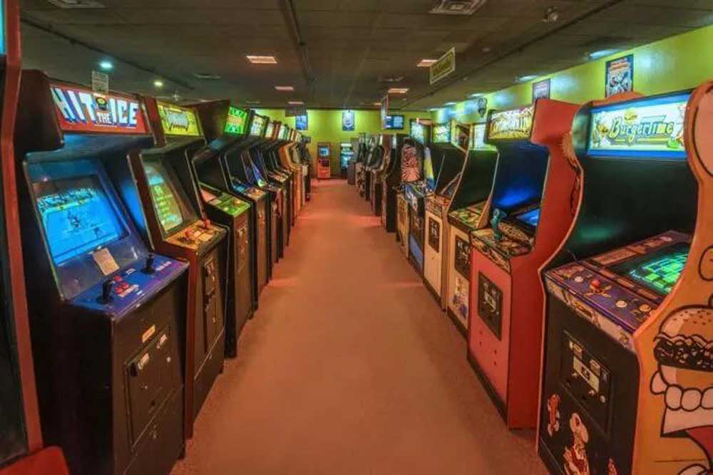 A brightly lit arcade with two rows of classic arcade game machines lining both sides of a carpeted walkway, creating a nostalgic, retro gaming atmosphere.