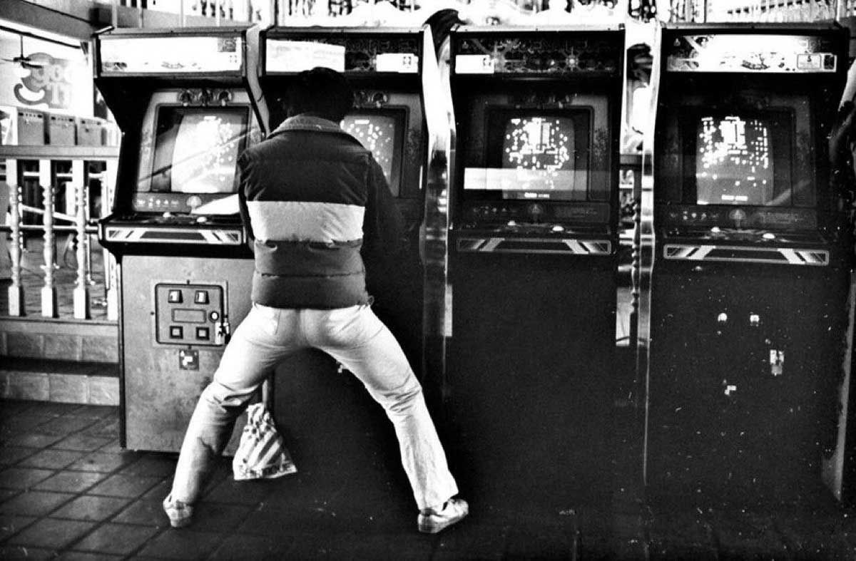 A person in a striped jacket and light pants stands with legs wide apart, playing on an arcade machine in a row of four vintage arcade games in a retro gaming area.