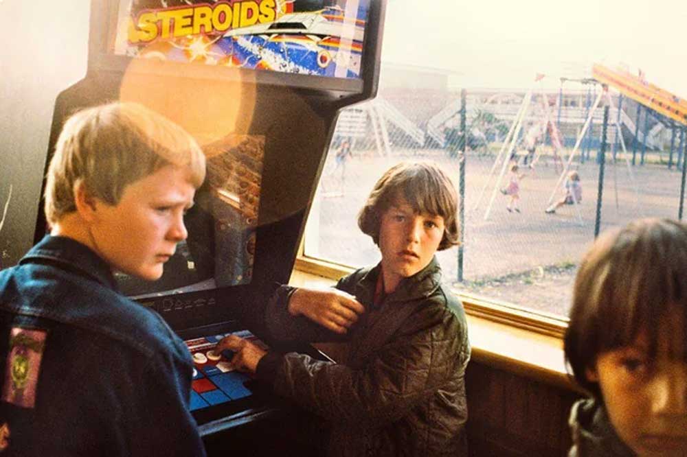 Three boys stand around a vintage arcade game labeled Steroids inside a building. Sunlight streams through a window, revealing a playground with children and swings outside. The boys look towards the camera.