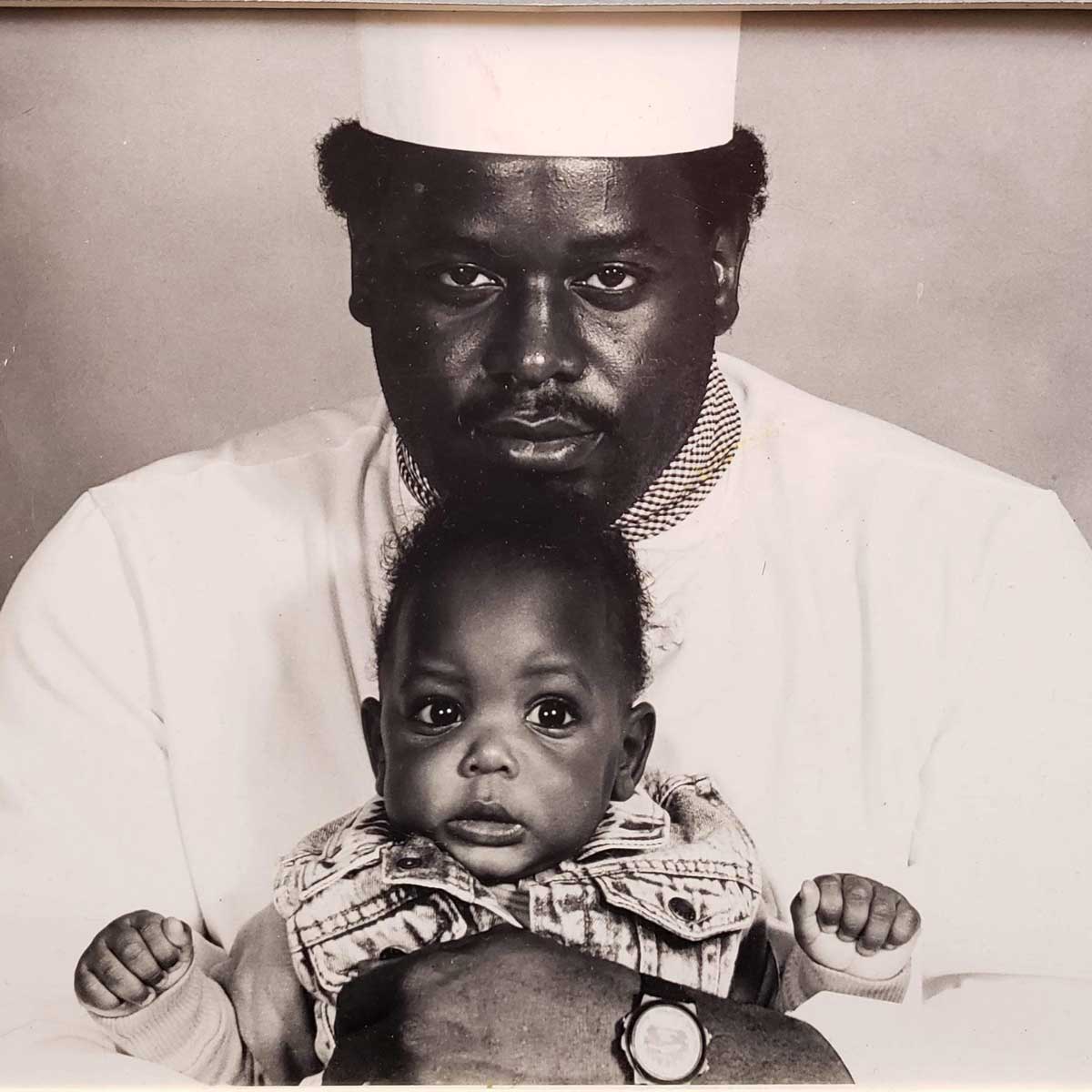 A Black man wearing a white hat and shirt holds a baby in his arms. The baby, dressed in a denim vest, looks toward the camera with wide eyes. The photo is in black and white.