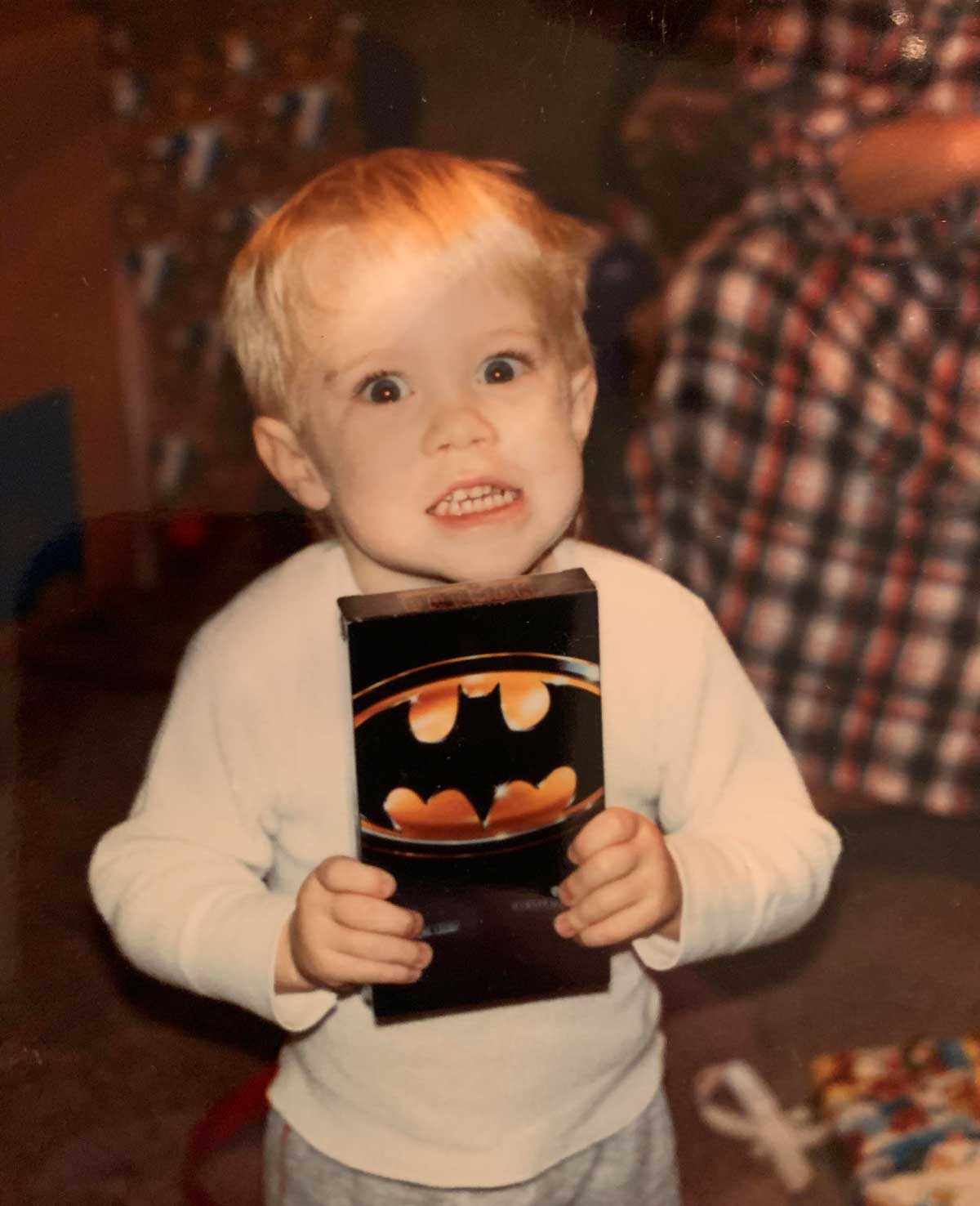 A young child with blond hair and a wide, excited expression holds a VHS tape with the Batman logo on the cover. The child wears a white shirt and stands indoors with blurred objects in the background.