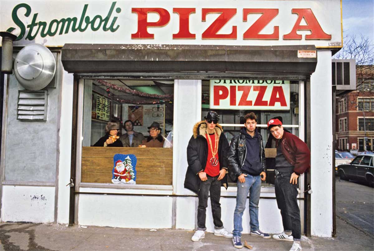 The Beastie Boys in casual 1980s attire stand outside Stromboli Pizza, a classic storefront with large windows. People sit inside eating, and a festive Santa decoration is visible on the window.