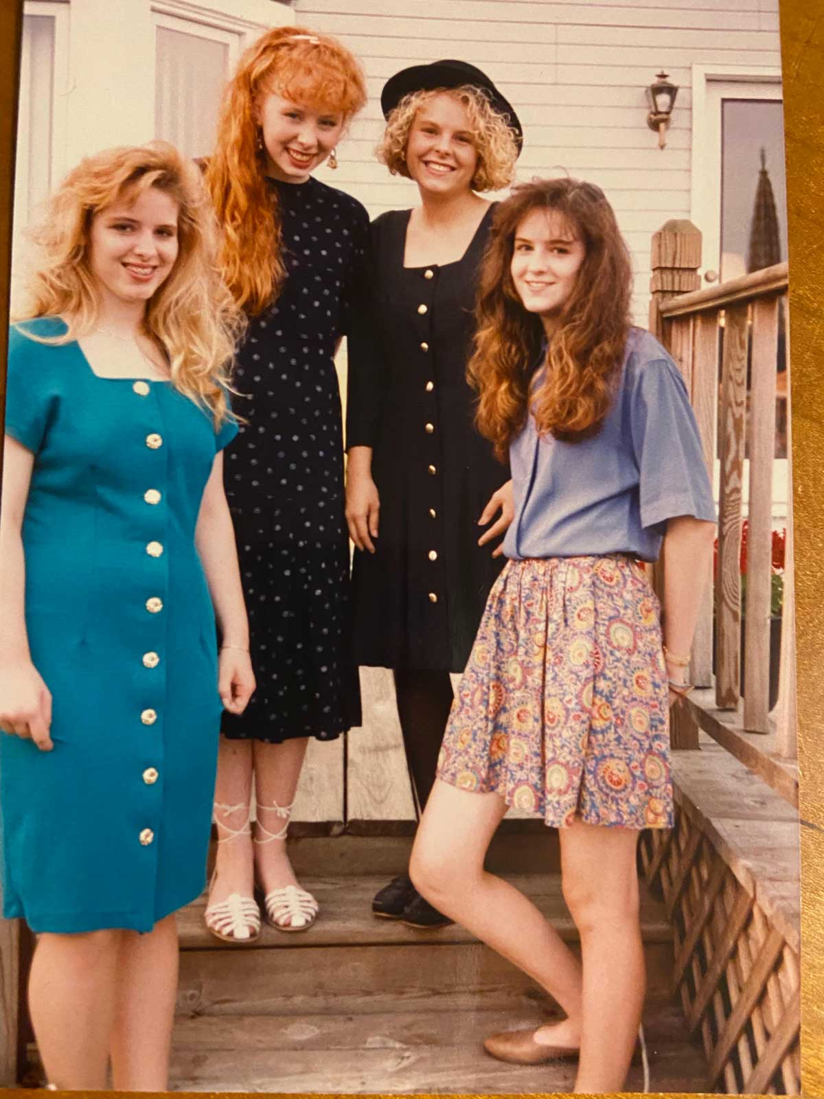 Four young women stand on outdoor steps, smiling at the camera. They wear colorful, vintage-style outfits, including dresses and skirts with buttons, and have various hairstyles, suggesting a 1990s or late 1980s fashion era.