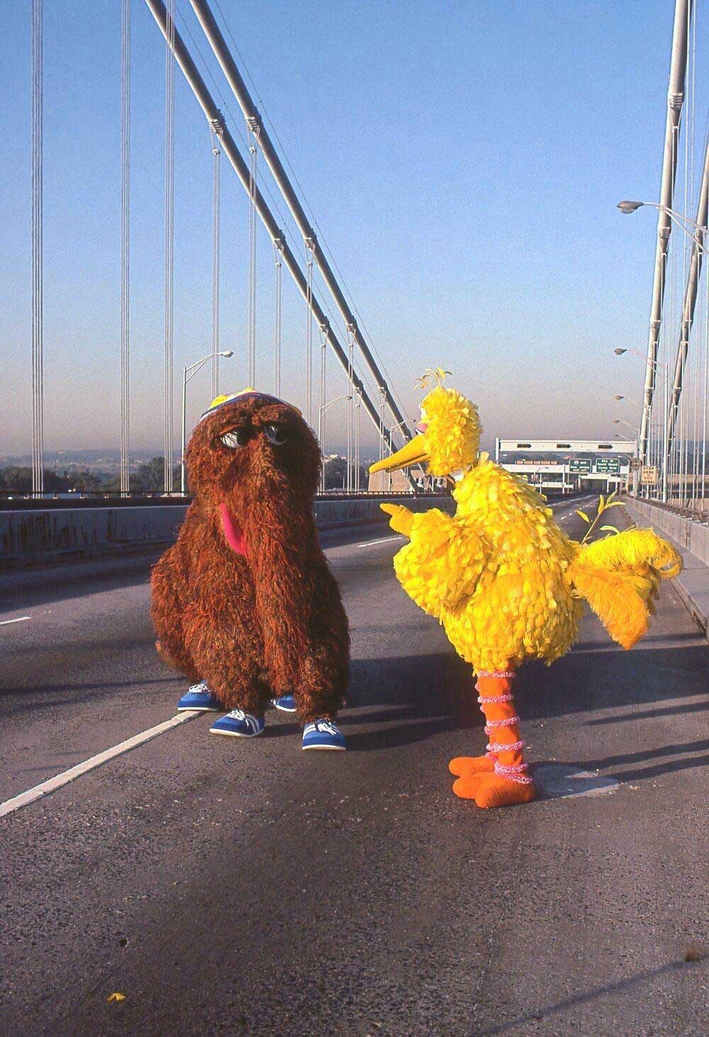 Two people in costumes—a large brown, furry creature and a tall, yellow bird—stand on an empty bridge under a clear blue sky. The scene is playful and whimsical.